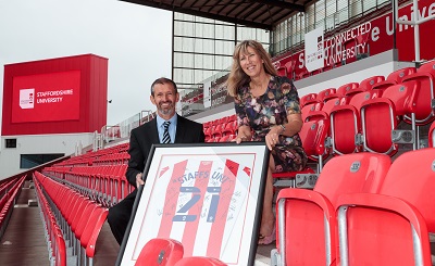 Man and woman pose in red stadium seats with a framed football shirt.