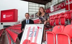 Man and woman pose in red stadium seats with a framed football shirt.