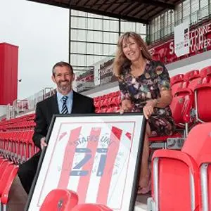 Man and woman pose in red stadium seats with a framed football shirt.