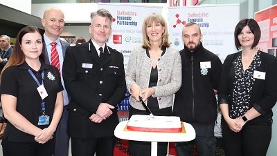Six people, including police officers, pose behind an anniversary cake at a forensic partnership event.