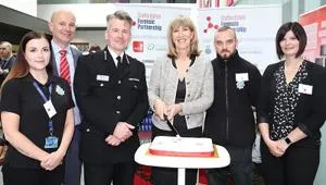 Six people, including police officers, pose behind an anniversary cake at a forensic partnership event.
