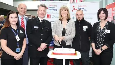 Six people, including police officers, pose behind an anniversary cake at a forensic partnership event.