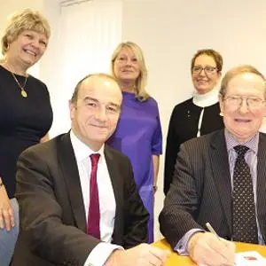 Two men sign a document at a table while three women stand behind them.