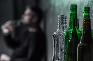 An image of alcohol bottles with a blurred figure drinking in the background