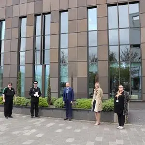 Six people stand spaced apart outside a modern brown-clad university building.