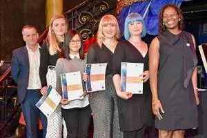 Five women at an awards ceremony hold certificates and printed programmes.