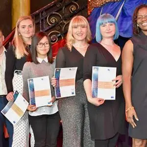 Five women at an awards ceremony hold certificates and printed programmes.