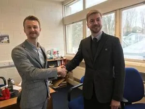Two men shake hands in a bright office, one wearing a suit and tie.