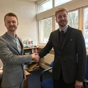 Two men shake hands in a bright office, one wearing a suit and tie.