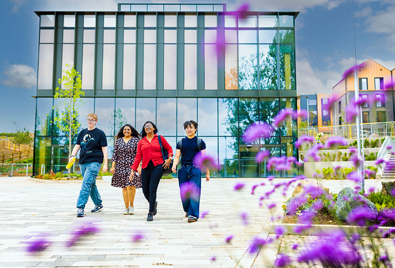 Four smiling students walking on front of a modern glass-fronted building. 