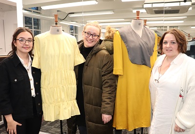 Three women stand beside mannequins dressed in cream and mustard sustainable garments.
