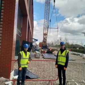 Two workers in hard hats and high-visibility vests stand at a large construction site.