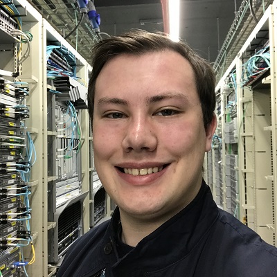 Smiling young man takes a selfie between rows of equipment in a server room.
