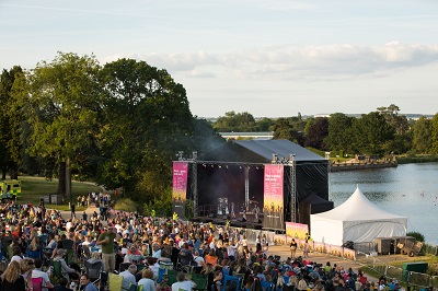 Large crowd watches an outdoor concert on a lakeside stage surrounded by trees.