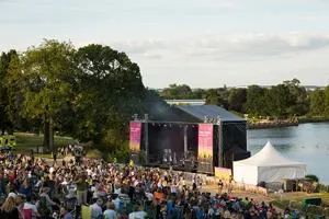 Large crowd watches an outdoor concert on a lakeside stage surrounded by trees.