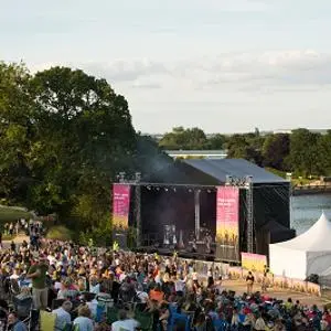 Large crowd watches an outdoor concert on a lakeside stage surrounded by trees.