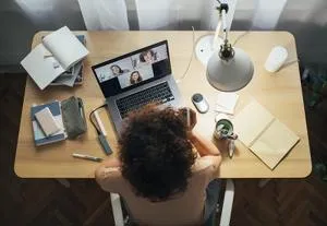 Overhead view of a desk with a video call on a laptop, notebook, phone, lamp and coffee.