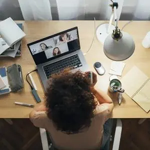 Overhead view of a desk with a video call on a laptop, notebook, phone, lamp and coffee.