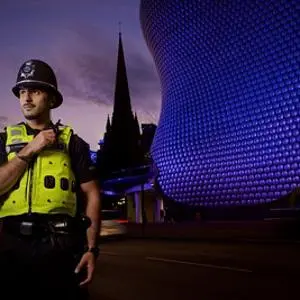 Police officer stands at night in Birmingham with the illuminated Bullring building behind.