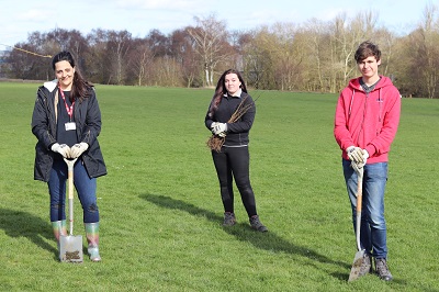 Three Staffordshire University students outdoors in wellington boots and warm clothing standing on grass with spades ready to dig.