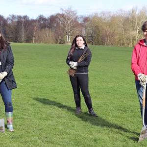Three Staffordshire University students outdoors in wellington boots and warm clothing standing on grass with spades ready to dig.