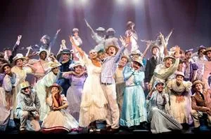 Large musical theatre cast in colourful costumes celebrates together on stage.