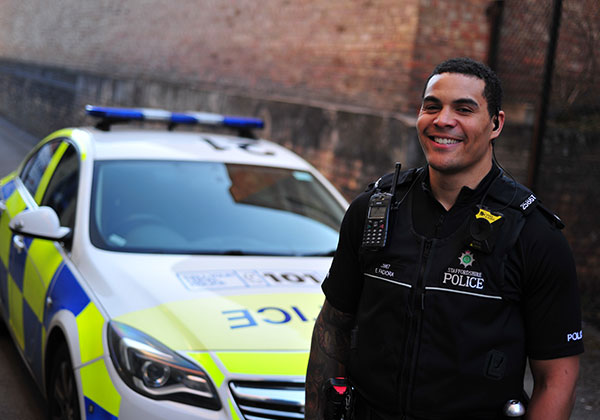 Image of a single male police officer smiling standing in front of a marker police car