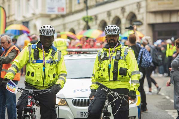 Image of two smiling police officers on bicyles in a crowded street with a police car in the background