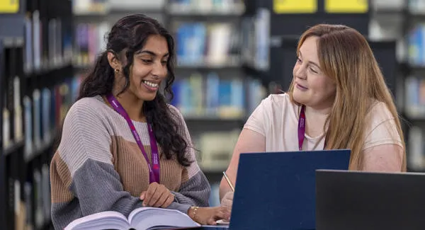 A female student with long dark hair and another female student with long blonde hair are studying together in the library with laptops
