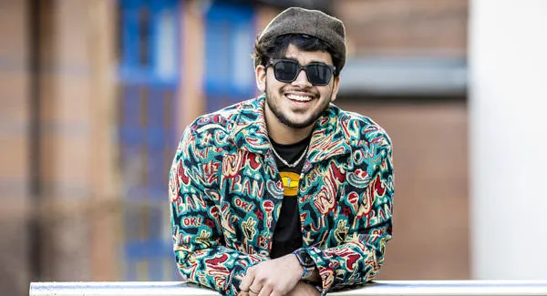 A male student wearing sunglasses and a hat is posing casually outside smiling
