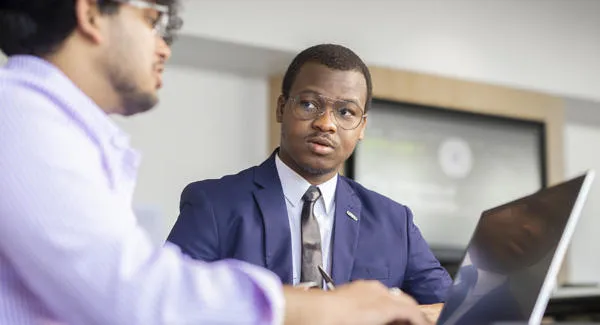 A young male student wearing a blue blazer shirt and tie is sitting at a desk talking to a smartly dressed man using a laptop