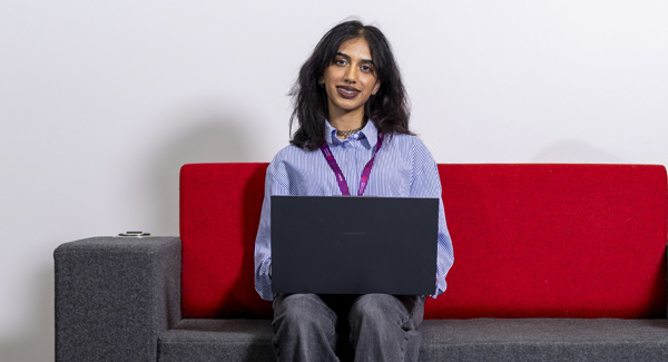 A female student with dark medium length hair wearing a blue shirt is smiling working on her laptop