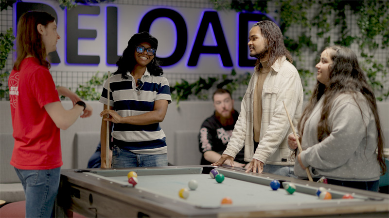 A group of young diverse students are playing pool in the break out area