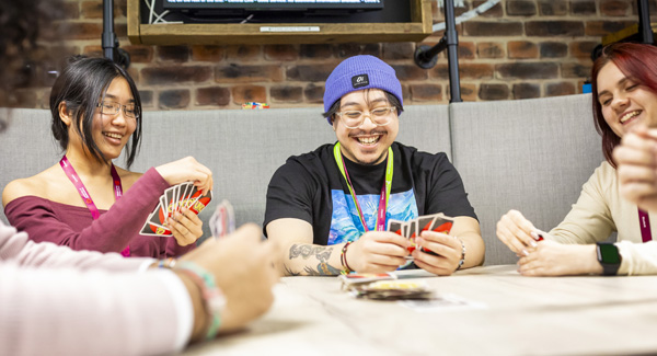 A group of casually dressed students are smiling whilst playing a card game in a modern breakout space