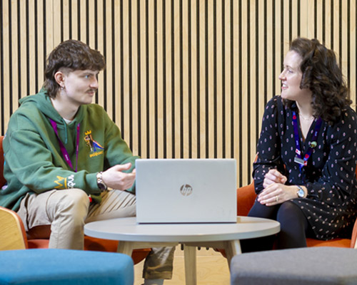 A casually dressed male student is chatting to an academic, they are sitting down in a modern open space with a laptop