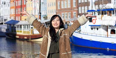 Young Asian woman wearing a brown coat stands with her arms outstretched in front of the waterfront in Nyhavn, Copenhagen