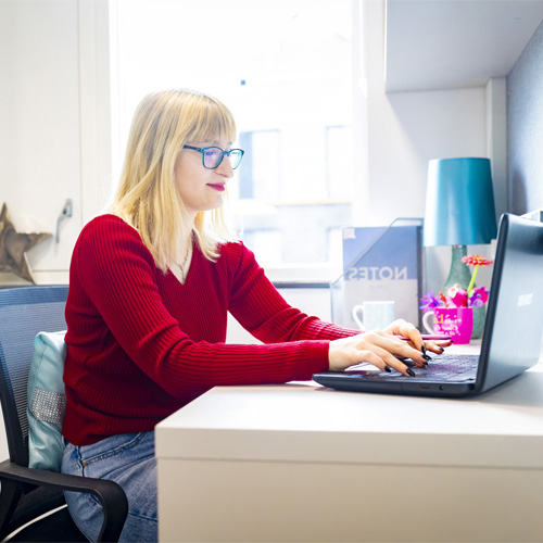 A female student with blonde hair is wearing a red jumper and using her laptop in a modern furnished student room