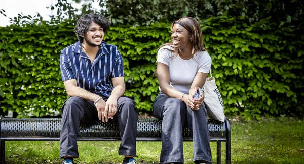 A male and a female student are smiling and sitting on a bench in front of a hedge near campus on a bright day