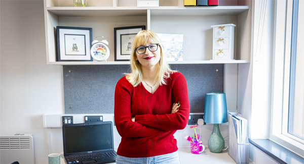 A female student with blonde hair, wearig a red jumper is standing near her desk in a modern student room