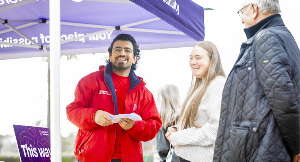 A student ambassador wearing a red uniform is chatting to a parent and student visitor at an open day