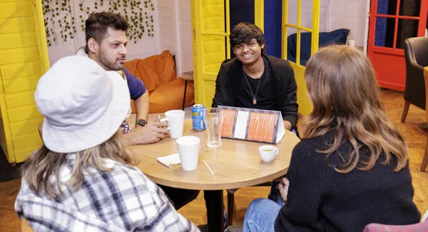 Studens are chatting at a table in the social space, there are colourful chill out zones and cushions around them