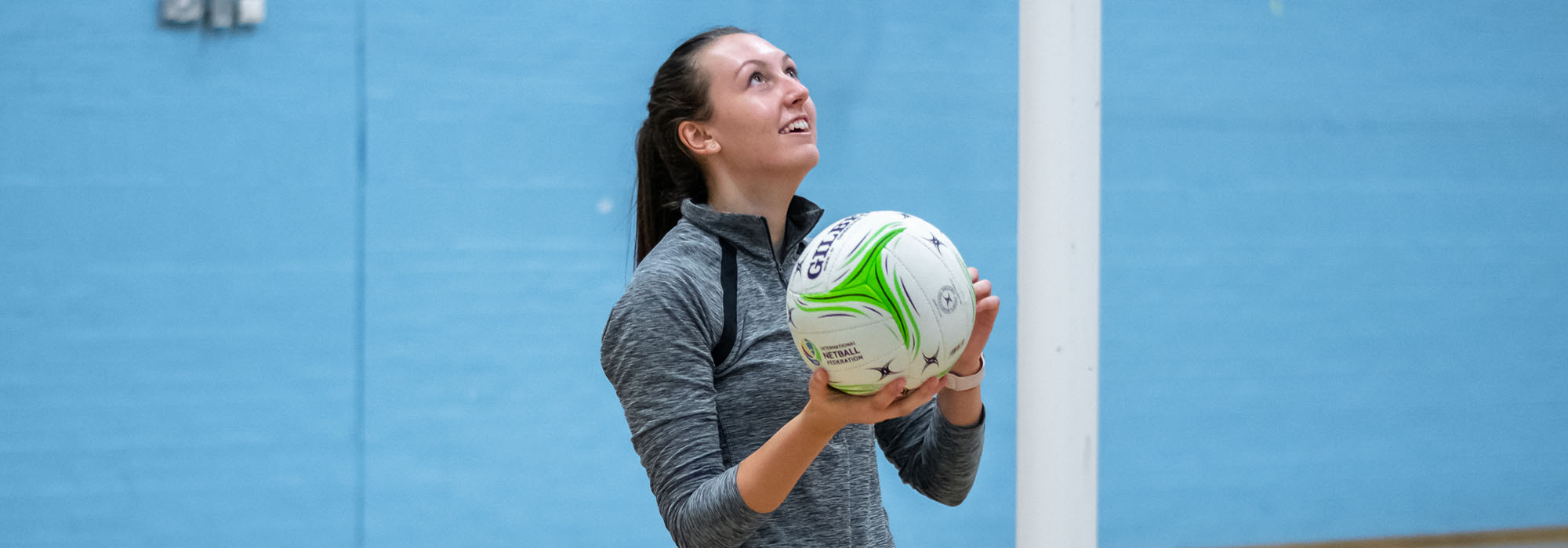 female netball player in the sports hall, holding a netball and looking up to the goal. she is wearing a grey top and has her long hair tied back,