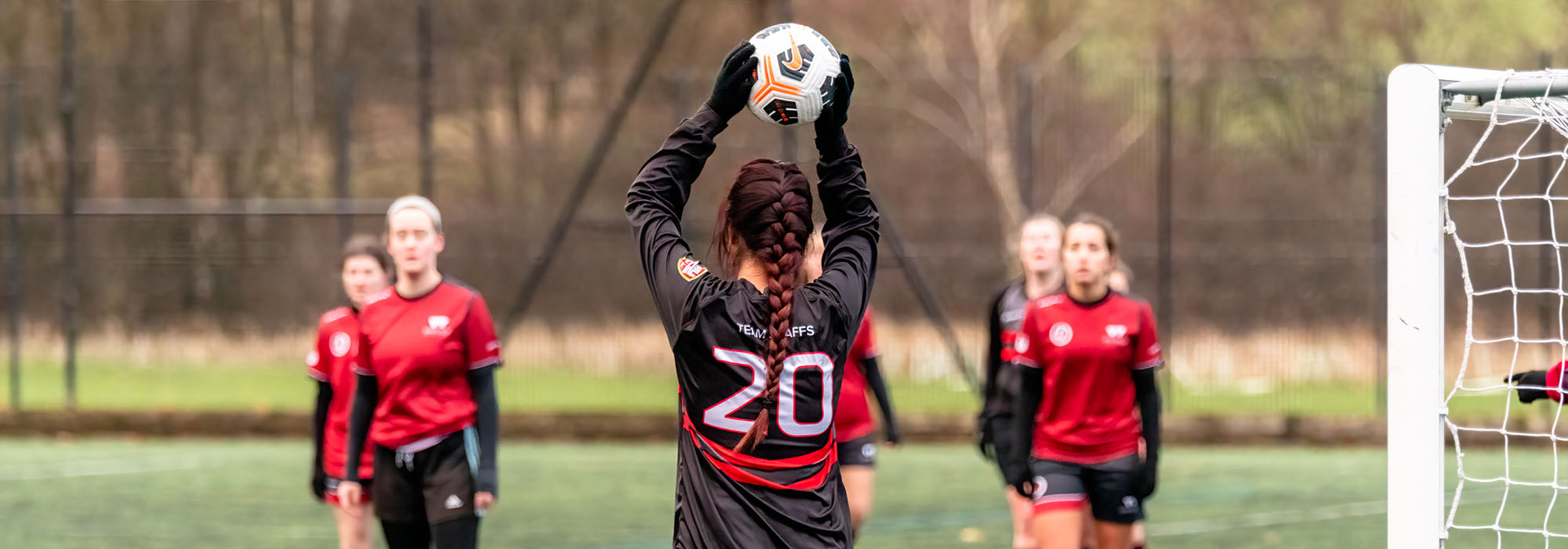 Staffs women's football player is standing on the side of the pitch and holds the ball above her head for a throw in. She has long hair in a plait and wears the Team Staffs kit.