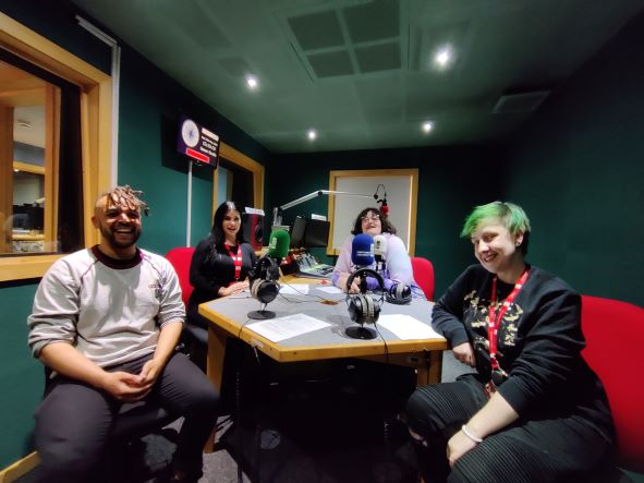 Mixed group of four students sitting around a desk in a recording studio with dark green walls. From left to right, Nesta, Holly, Alex and Maxie all smile to the camera. They are dressed casually..