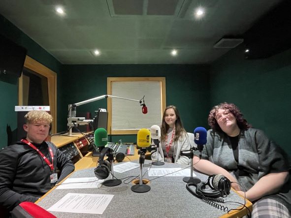 Mixed group of four students sitting around a desk in a recording studio with dark green walls. From left to right, Danny, Tiff and Alex all smile to the camera. They are dressed casually.