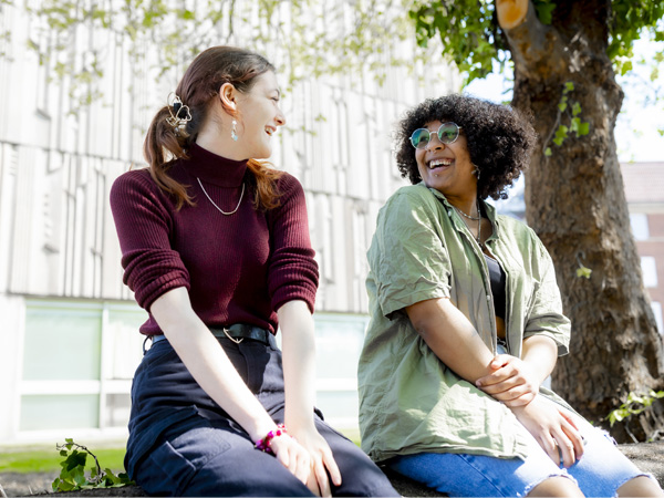 Two female students dressed casually, are sitting next to a tree, they are smiling and chatting to one another