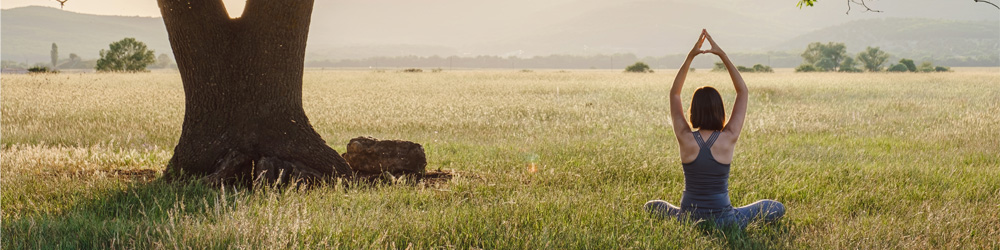 Image of a woman wearing yoga clothes, sitting crosslegged with her arms stretched above her head, in a field close to a large tree and looking out towards a sunset