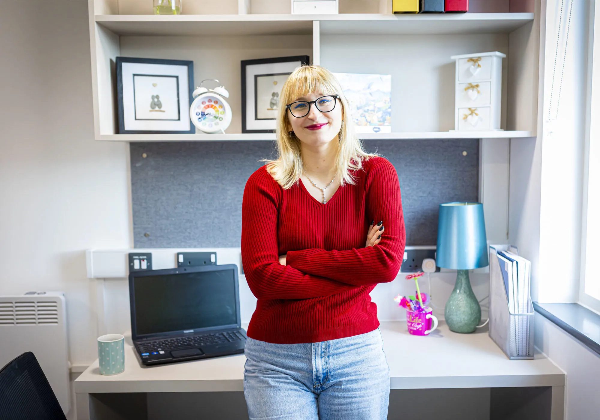 A female student with blonde hair is standing infront of her desk in student accommodation