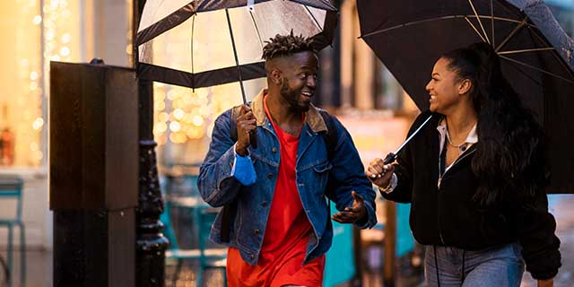 male and female student walk down high street, they are holding umbrellas