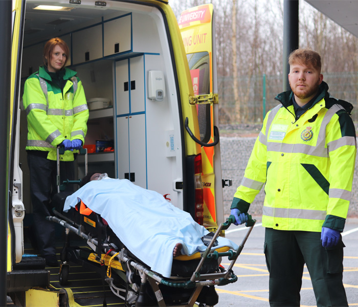 Paramedic students are dressed in uniform practising a health simulation with an emergency ambulance and patient